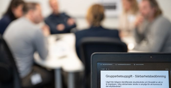 Datorskärm i förgrunden och en grupp kursdeltagare i bakgrunden.