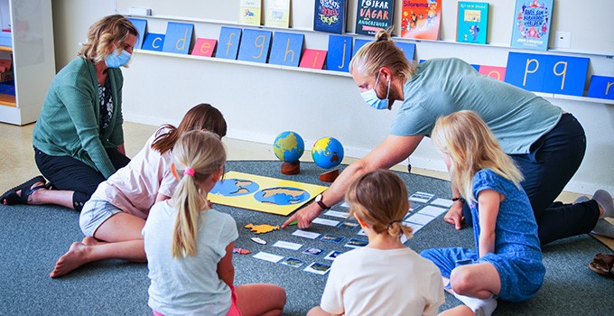 Bild från skola där fyra elever sitter på en matta och spelar spel tillsammans med två pedagoger som bär munskydd. 