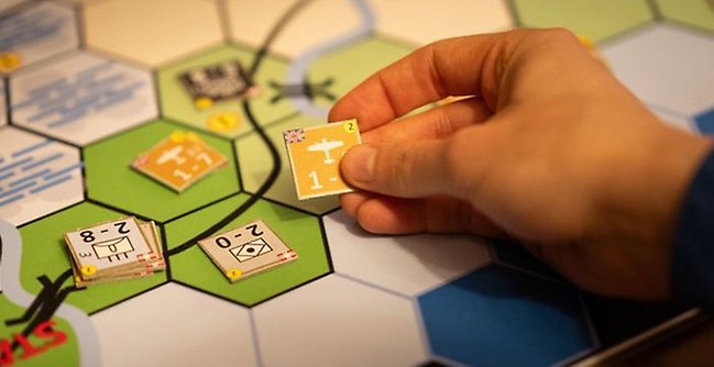 Hand som håller i en spelbricka över ett brädspel. 