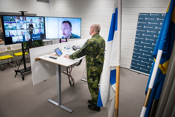 Military officer standing in front of two big screens with twelve people joining meeting digitally.