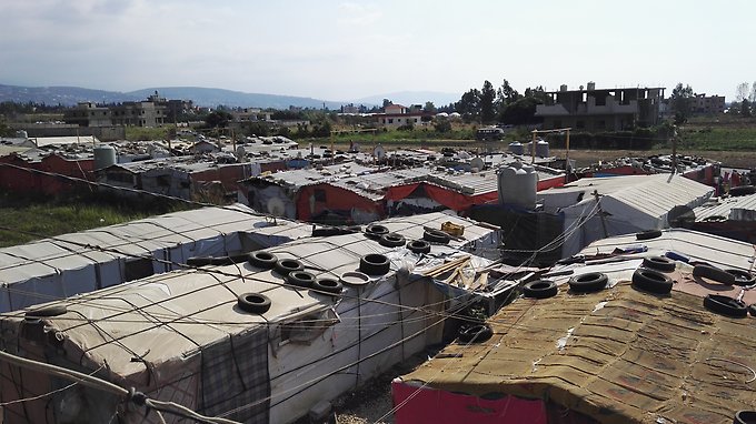 Flyktingläger med flera stora tält på rad. I bakgrunden finns bostadshus och i fjärran berg.