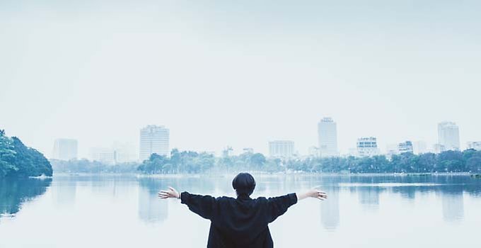 Kvinna som håller upp armarna framför sjö med stadsmotiv i bakgrunden.